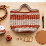 Bolsa de crochê artesanal estruturada com fio de malha premium terracota sobre mesa de madeira, acompanhada de alça de couro sintético, ferragens douradas com mosquetões, fecho magnético, base de couro e agulha ergonômica.