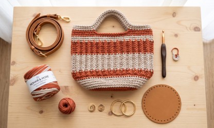 Bolsa de crochê artesanal estruturada com fio de malha premium terracota sobre mesa de madeira, acompanhada de alça de couro sintético, ferragens douradas com mosquetões, fecho magnético, base de couro e agulha ergonômica.
