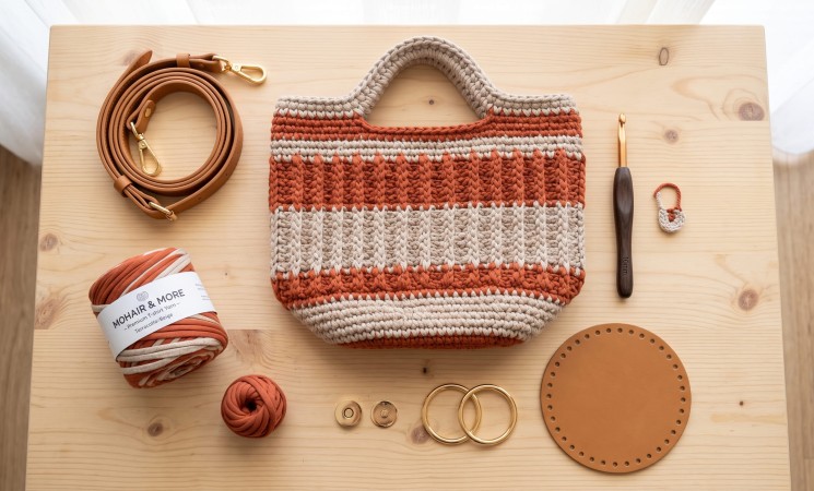 Bolsa de crochê artesanal estruturada com fio de malha premium terracota sobre mesa de madeira, acompanhada de alça de couro sintético, ferragens douradas com mosquetões, fecho magnético, base de couro e agulha ergonômica.
