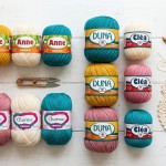 Novelos de linha de crochê Círculo das marcas Anne, Charme, Duna e Cléa organizados em paleta de cores rosa, amarelo, cru e azul-petróleo ao lado de agulhas de bambu e amostra rendada sobre mesa branca.