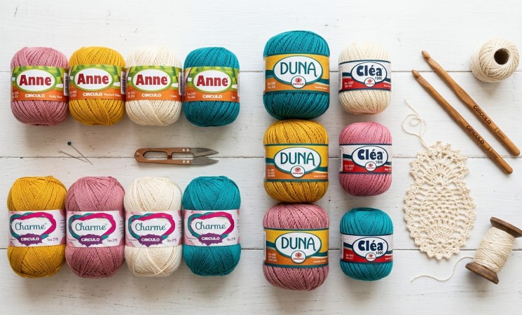 Novelos de linha de crochê Círculo das marcas Anne, Charme, Duna e Cléa organizados em paleta de cores rosa, amarelo, cru e azul-petróleo ao lado de agulhas de bambu e amostra rendada sobre mesa branca.